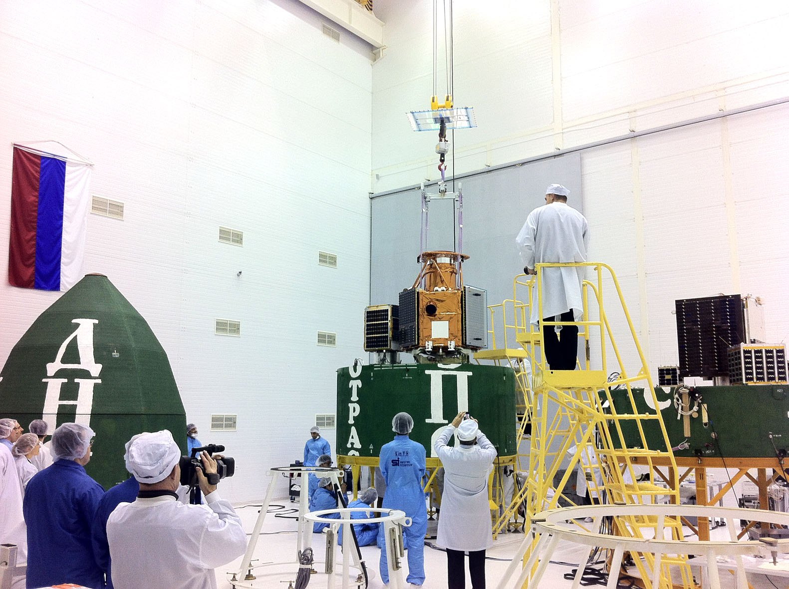 Technicians in clean-room suits working around a cylindrical rocket payload module holding several satellites
