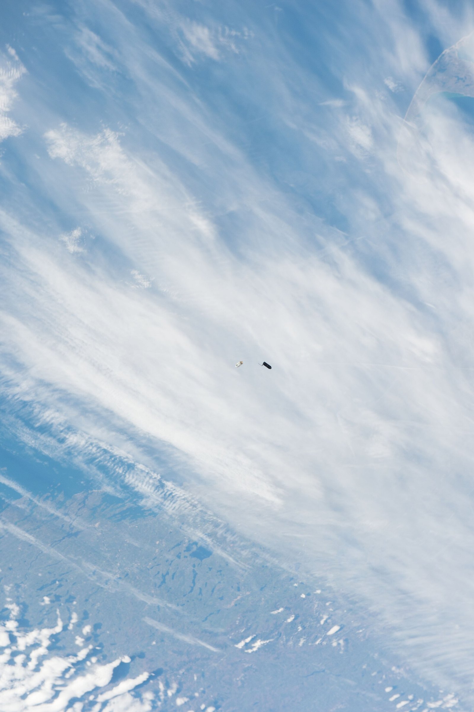 Two small box-shaped Dove CubeSats with solar panels drifting away from the International Space Station above a cloudy ocean