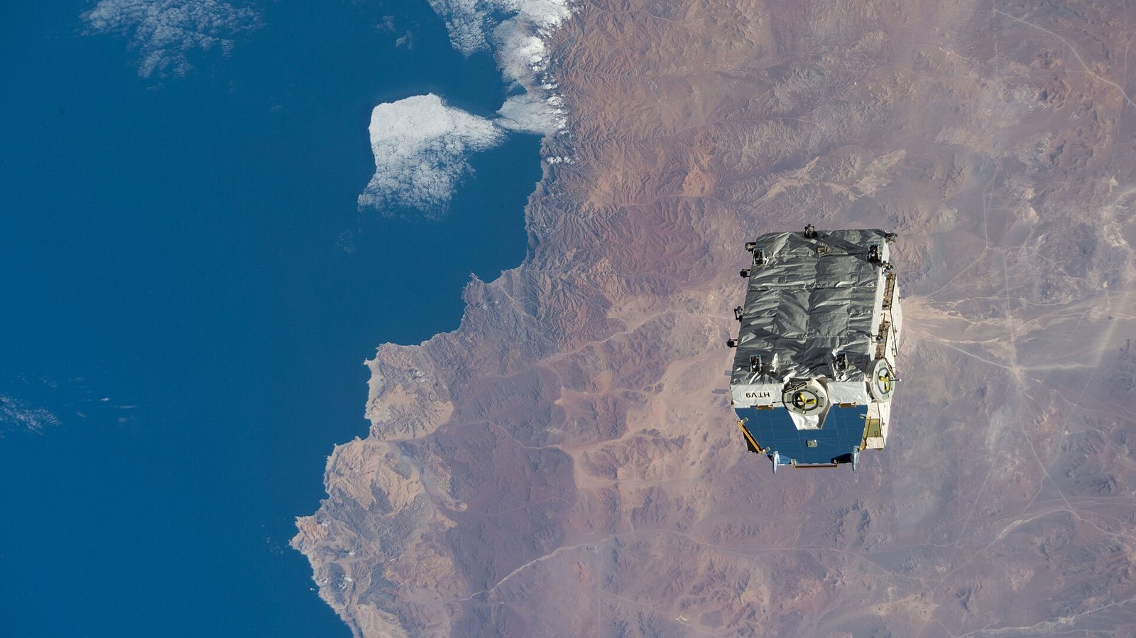 A pallet loaded with cylindrical battery canisters drifting away from the International Space Station above a cloud-covered ocean