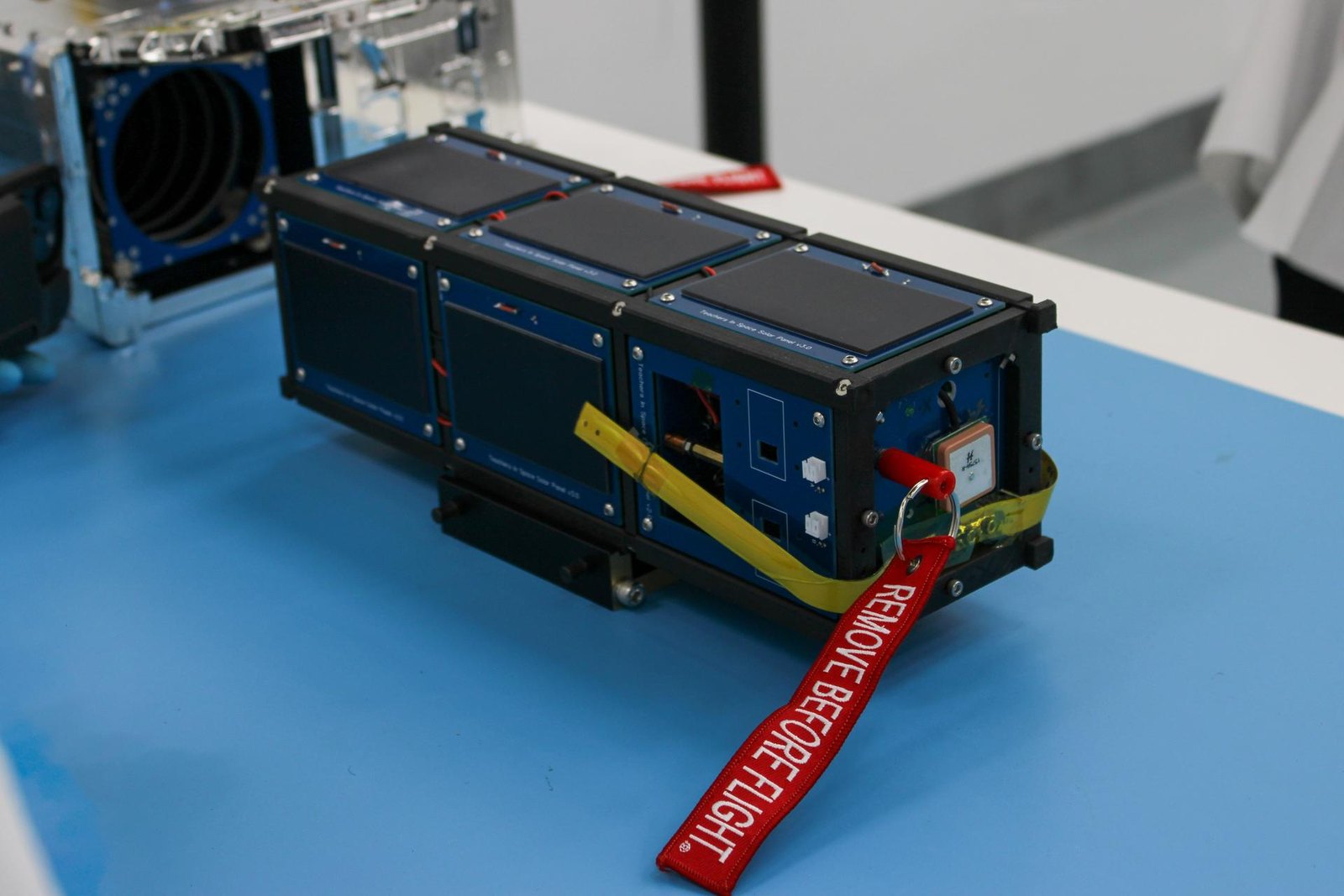 A small three-unit CubeSat standing on a bench in a payload processing clean room