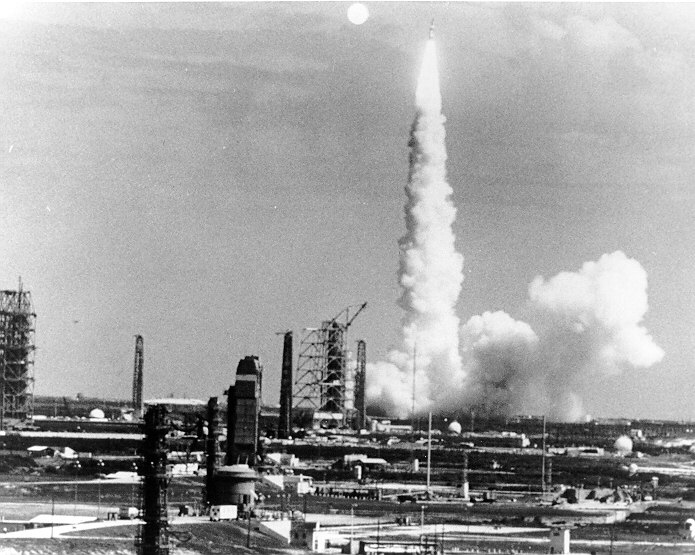 A Titan IIIC rocket climbing away from its launch pad trailing a bright exhaust plume against a pale sky