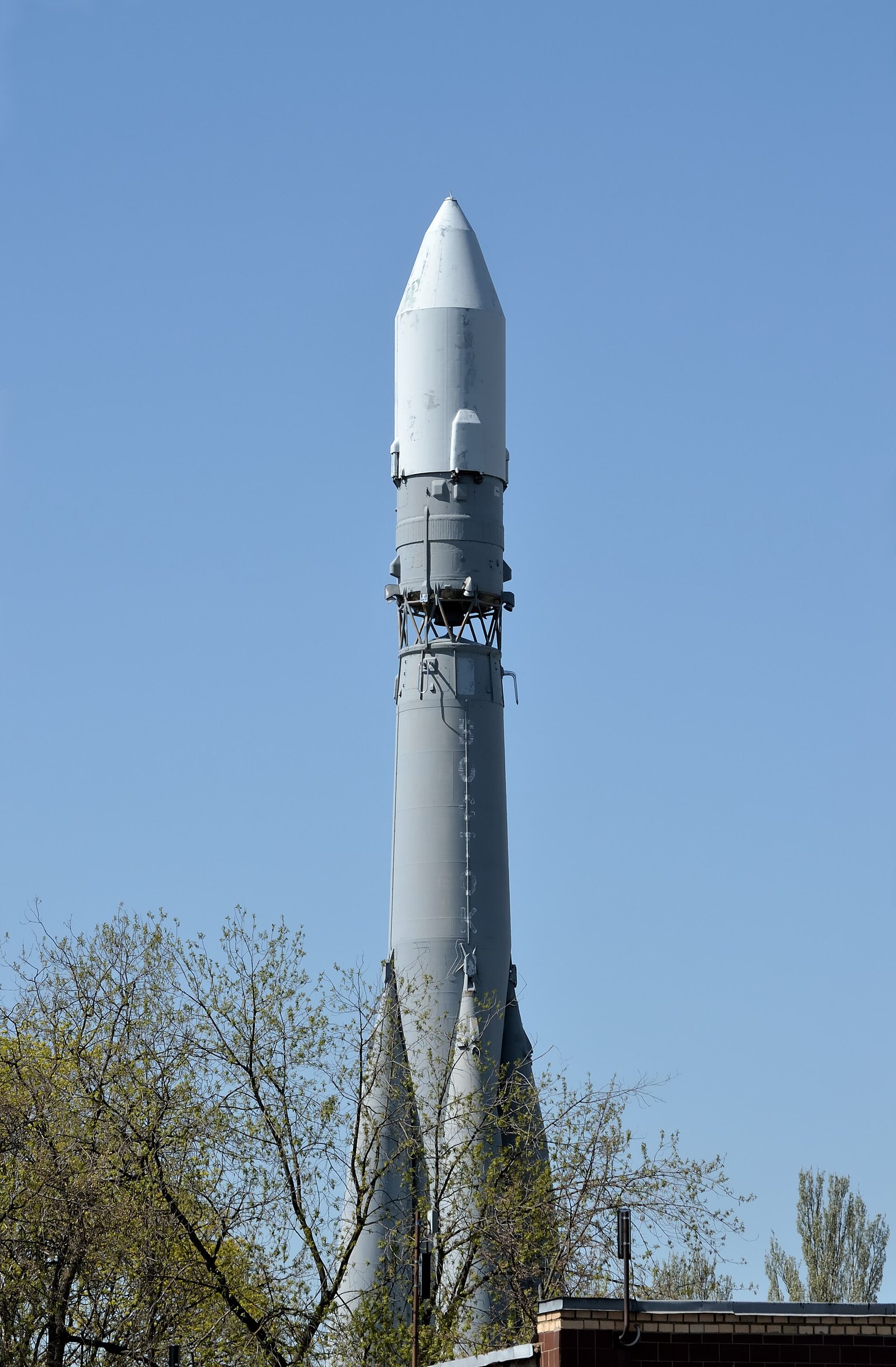 A full-scale model of the Vostok-2M carrier rocket standing upright as a monument on a paved square, with trees and a building behind it