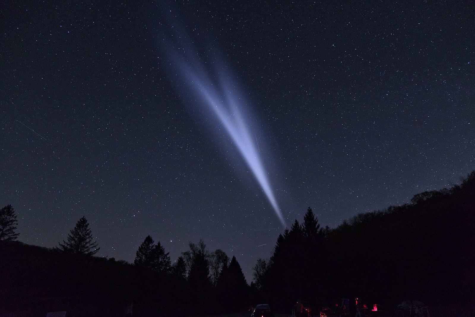 A faint luminous cloud of vented rocket propellant spreading across a dark starry sky above a field, with red equipment lights at lower right